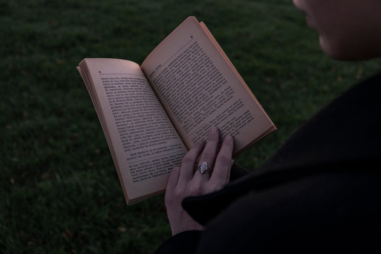 Person holding and reading a book outdoors during twilight, focus on hands and open pages.